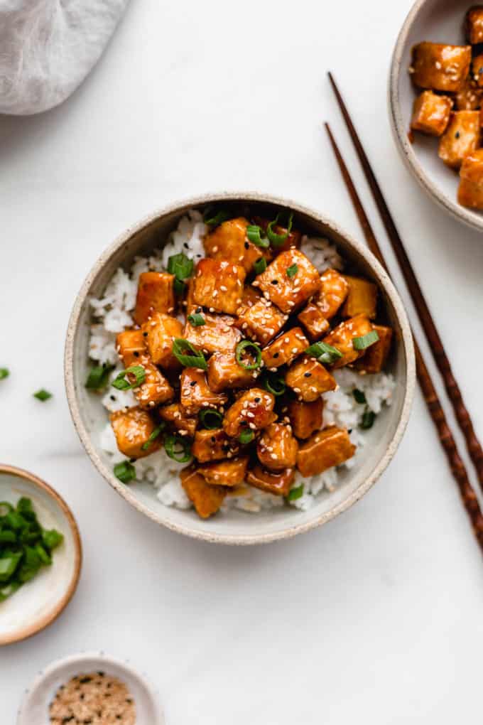 Sweet and sour tofu in a white ceramic bowl with two small bowls on the side filled with green onions and sesame seeds